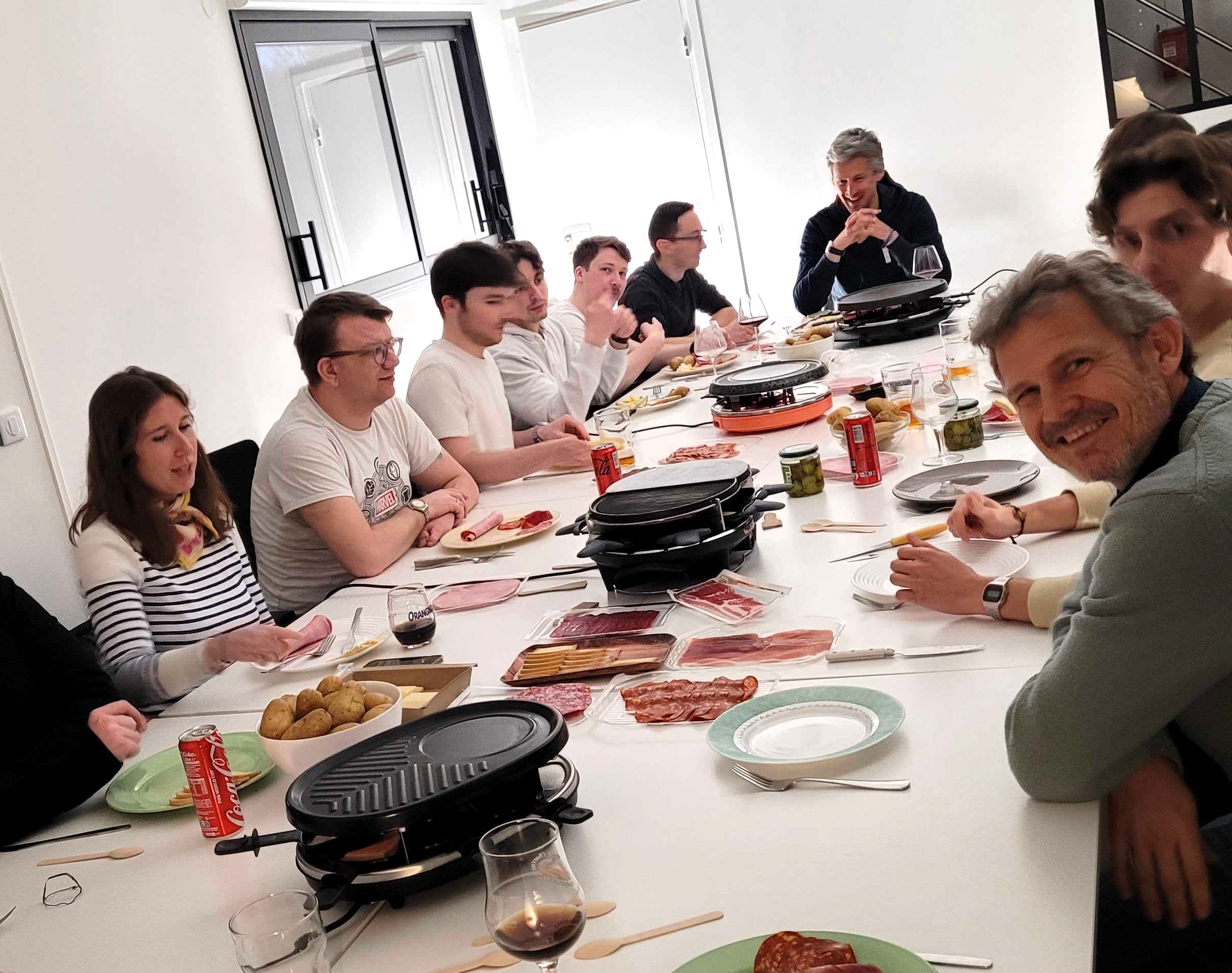 Équipe IT-Room réunie autour d'une table avec appareil à raclette et charcuterie, lors d'un moment convivial entre collègues